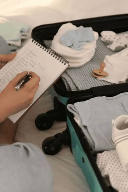 Une future maman prépare sa valise de maternité sur un canapé, en cochant une liste manuscrite à côté d'un ordinateur portable affichant le site d'Elsan avec des vêtements et accessoires pour bébé.
