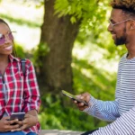 Deux jeunes adultes sourient en se parlant, téléphone à la main, assis dans un parc | labonnecopine.com