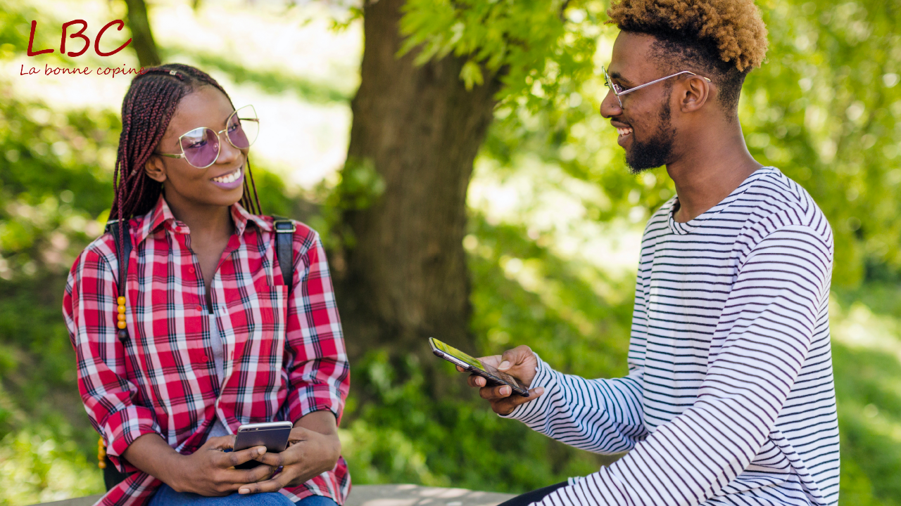 Deux jeunes adultes sourient en se parlant, téléphone à la main, assis dans un parc | labonnecopine.com