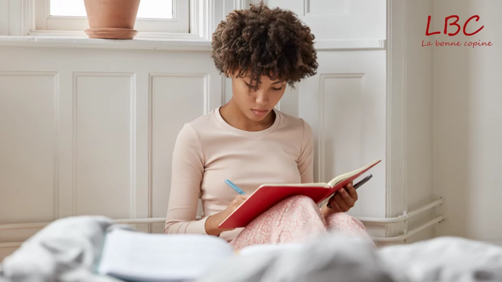Assise près de la fenêtre, le regard apaisé, une femme écrit dans un carnet rouge. Chaque mot posé est une étape de plus vers elle-même, un moment de recentrage, de clarté, de reconnexion | labonnecopine.com