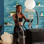 Une femme en pleine séance d’entraînement sur un vélo elliptique, installée dans un salon au design moderne. Une scène qui allie bien-être, motivation et confort à domicile | labonnecopine.com