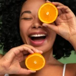 Une femme souriante tient fièrement une demi-orange juteuse, contrastant joliment avec un décor aux tons verts éclatants. Son expression rayonnante et la scène colorée évoquent la vitalité, la fraîcheur et le plaisir d’adopter une routine saine et naturelle | labonnecopine.com