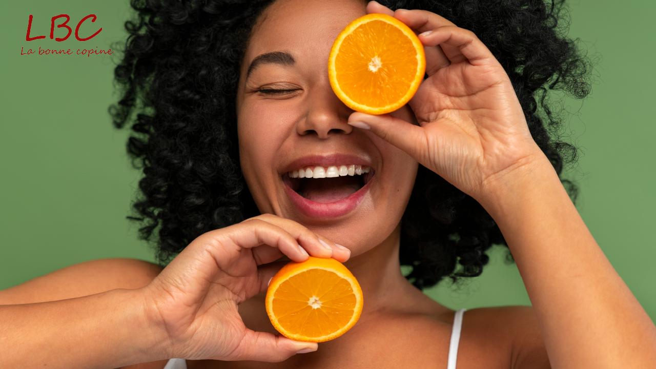Une femme souriante tient fièrement une demi-orange juteuse, contrastant joliment avec un décor aux tons verts éclatants. Son expression rayonnante et la scène colorée évoquent la vitalité, la fraîcheur et le plaisir d’adopter une routine saine et naturelle | labonnecopine.com