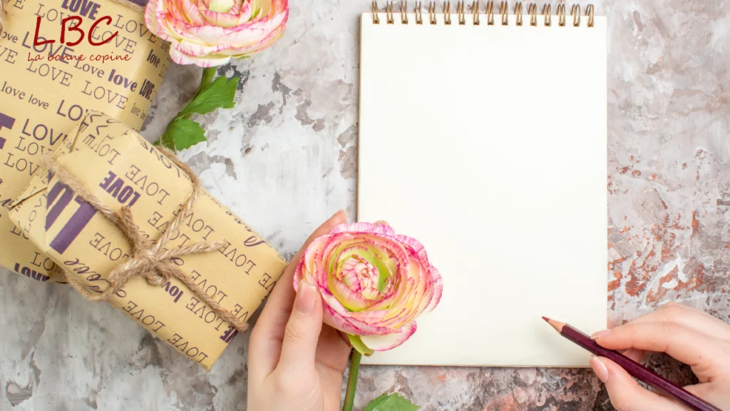 Entre douceur et intention, une main enlace une fleur et un crayon, prête à poser des mots sur un carnet vide. À ses côtés, des présents emballés évoquent le soin et la générosité | labonnecopine.com