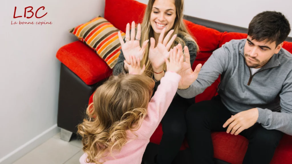 Un enfant, tout sourire, tend les mains pour donner des &laquo; high five &raquo; &agrave; deux adultes assis confortablement sur un canap&eacute; rouge, une sc&egrave;ne pleine de vie et de bonne humeur | labonnecopine.com