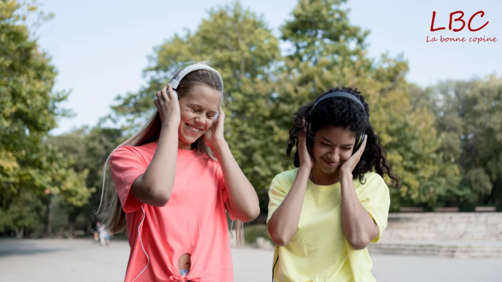 Dans un parc baigné de verdure, deux jeunes femmes partagent un moment complice en écoutant de la musique, sourires aux lèvres. Une scène simple et joyeuse qui illustre la convivialité et le pouvoir rassembleur du son | labonnecopine.com
