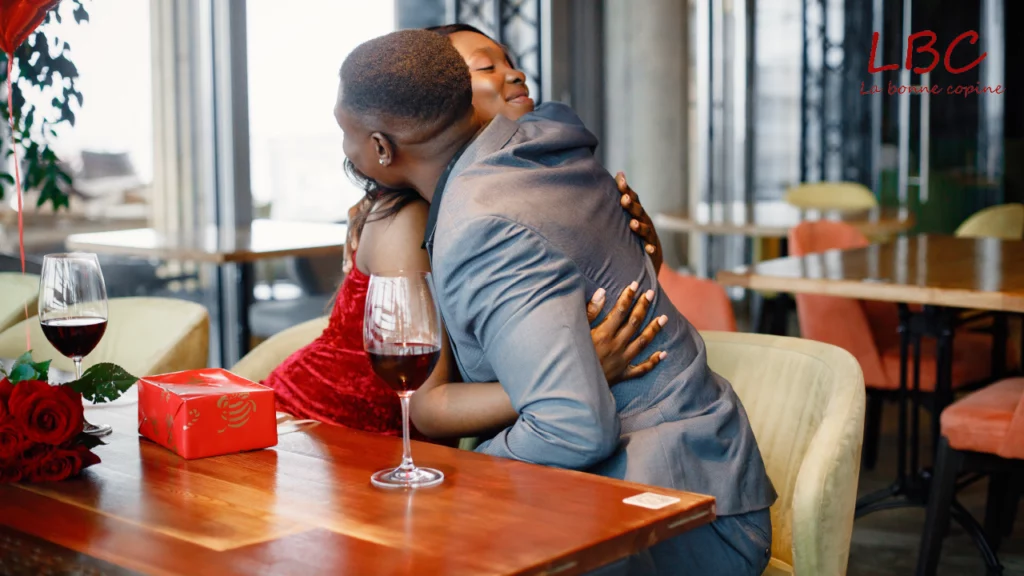Texte alternatif :
Un couple s'enlace tendrement dans un restaurant élégant. L'homme en veste grise étreint une femme en robe rouge velours. Sur leur table en bois se trouvent deux verres de vin rouge, un petit cadeau emballé et quelques roses | labonnecopine.com