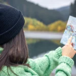 Sous un ciel clair, une femme portant un bonnet noir et un pull vert scrute attentivement une carte aux trajets multicolores. Devant elle, un paysage grandiose s’étend : un lac scintille au pied de montagnes imposantes, dessinant une scène à la fois paisible et inspirante | labonnecopine.com