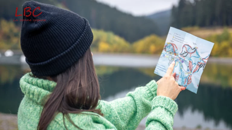 Sous un ciel clair, une femme portant un bonnet noir et un pull vert scrute attentivement une carte aux trajets multicolores. Devant elle, un paysage grandiose s’étend : un lac scintille au pied de montagnes imposantes, dessinant une scène à la fois paisible et inspirante | labonnecopine.com