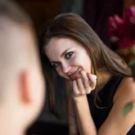 Une femme aux cheveux châtains adresse un sourire complice à une personne hors champ, tandis qu’un bouquet de fleurs rouges apporte une touche de chaleur en arrière-plan. Une scène empreinte de douceur et de connexion | labonnecopine.com