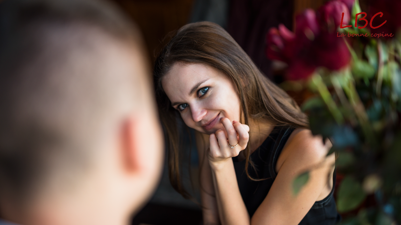 Une femme aux cheveux châtains adresse un sourire complice à une personne hors champ, tandis qu’un bouquet de fleurs rouges apporte une touche de chaleur en arrière-plan. Une scène empreinte de douceur et de connexion | labonnecopine.com