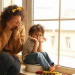 Une femme assise sur un rebord de fenêtre se couvre le visage, visiblement épuisée. À ses côtés, un petit garçon l’imite avec curiosité, entouré de jouets | labonnecopine.com