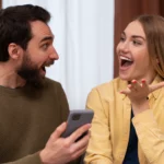 Installé sur un canapé, un couple échange avec le sourire autour d’un téléphone. Leurs gestes et regards complices reflètent la vitalité d’une communication authentique | labonnecopine.com