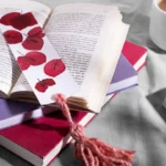 Une pile de livres, certains ouverts, d'autres fermés, est posée sur une table aux côtés d’une tasse de café. Sur l’un des ouvrages ouverts repose un marque-page orné de feuilles rouges et terminé par un pompon rose | labonnecopine.com