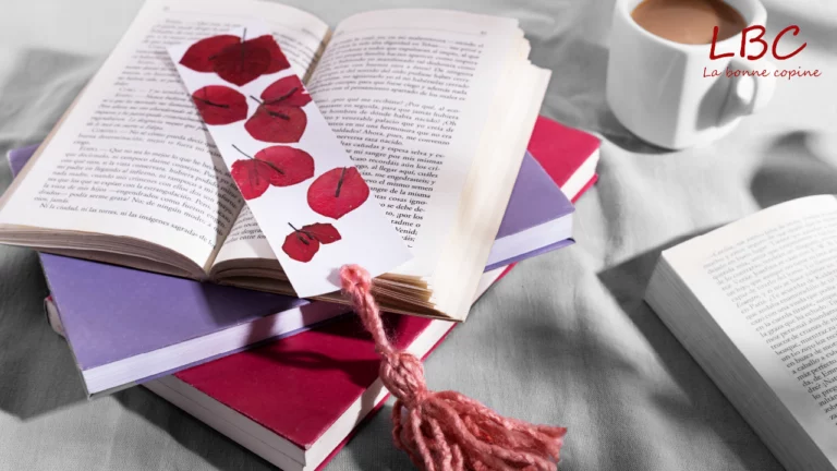 Une pile de livres, certains ouverts, d'autres fermés, est posée sur une table aux côtés d’une tasse de café. Sur l’un des ouvrages ouverts repose un marque-page orné de feuilles rouges et terminé par un pompon rose | labonnecopine.com