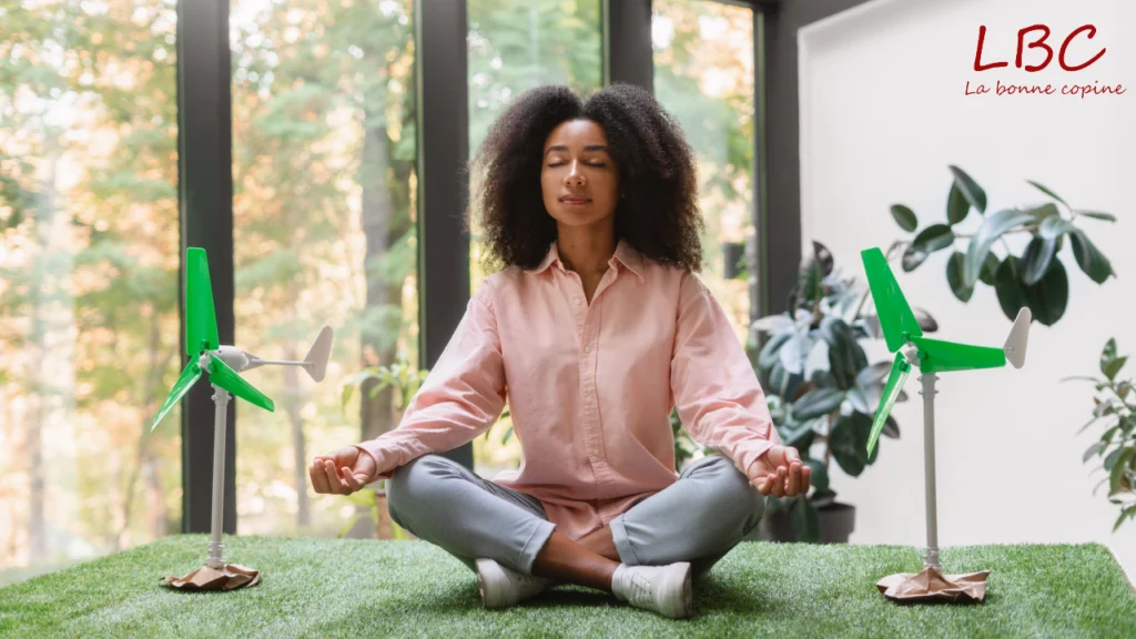 Dans un intérieur baigné de lumière naturelle, une femme médite en tailleur sur un tapis de gazon artificiel. Autour d’elle, de petites éoliennes vertes et des plantes d’intérieur créent une atmosphère apaisante, mêlant harmonieusement nature, énergie renouvelable et bien-être | labonnecopine.com