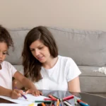 Assises côte à côte à une table dans un salon au style moderne, une femme guide doucement une petite fille qui dessine avec des crayons de couleur. Ensemble, elles partagent un moment simple et chaleureux, fait d’apprentissage et de créativité | labonnecopine.com