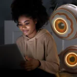 Une jeune femme souriante pose aux côtés de deux robots au design futuriste, reconnaissables à leurs yeux lumineux. La scène se déroule dans un environnement moderne, illustrant l’harmonie entre technologie et quotidien | labonnecopine.com