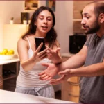Une femme montre son téléphone à un homme tout en lui parlant de manière expressive ; ils semblent en plein désaccord dans une cuisine moderne | labonnecopine.com