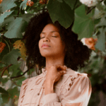 Dans un cadre naturel vibrant de vie, une femme se tient debout, entourée d’un mélange généreux de fleurs colorées et de feuillages abondants. Sa posture paisible et son expression sereine évoquent un profond sentiment de connexion à la nature, illustrant un moment d’équilibre et de ressourcement au cœur d’un environnement végétal apaisant | labonnecopine.com