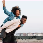 En pleine nature, un jeune homme transporte sur son dos une femme éclatante de sourire. Le couple, visiblement complice, partage un moment de bonheur simple et authentique, leur joie contagieuse illuminant la scène | labonnecopine.com