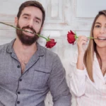 Un duo complice partage un moment de gaieté, chacun mordant délicatement la tige d'une rose écarlate. Lui, arborant une barbe soignée et vêtu d'une élégante chemise grise, affiche un sourire radieux. Elle, avec sa chevelure soyeuse cascadant sur ses épaules, porte un chemisier blanc à motifs subtils qui illumine son visage enjoué. Tous deux se détachent sur un fond immaculé aux textures boisées, créant une atmosphère à la fois intime et lumineuse | labonnecopine.com