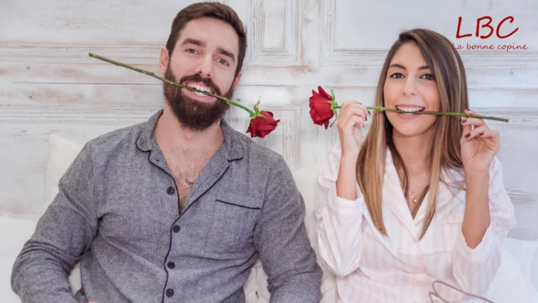 Un duo complice partage un moment de gaieté, chacun mordant délicatement la tige d'une rose écarlate. Lui, arborant une barbe soignée et vêtu d'une élégante chemise grise, affiche un sourire radieux. Elle, avec sa chevelure soyeuse cascadant sur ses épaules, porte un chemisier blanc à motifs subtils qui illumine son visage enjoué. Tous deux se détachent sur un fond immaculé aux textures boisées, créant une atmosphère à la fois intime et lumineuse | labonnecopine.com