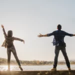 Dans la lumière dorée du soir, un couple s’élève bras ouverts devant un lac paisible, symbole d’harmonie, de légèreté et de bonheur partagé | labonnecopine.com