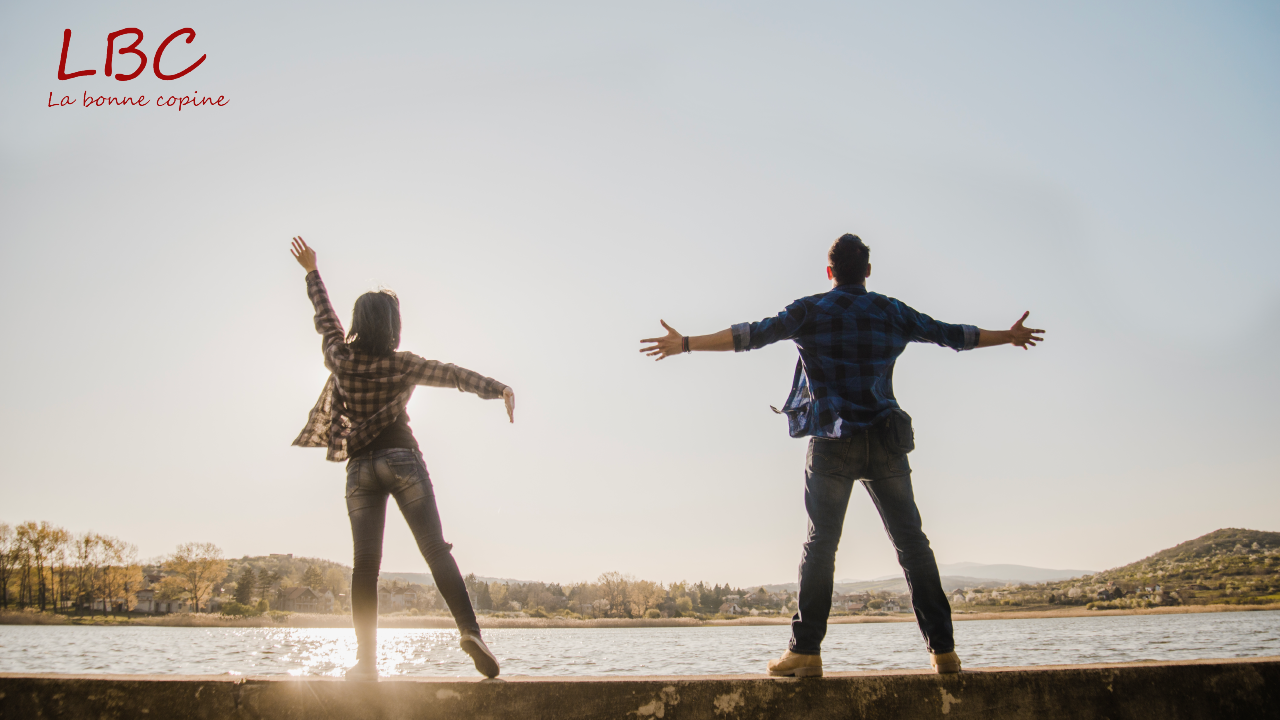 Dans la lumière dorée du soir, un couple s’élève bras ouverts devant un lac paisible, symbole d’harmonie, de légèreté et de bonheur partagé | labonnecopine.com