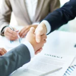 Deux personnes en costume échangent une poignée de main au-dessus d’un contrat fraîchement signé, symbolisant un accord conclu et une collaboration professionnelle scellée | labonnecopine.com