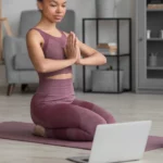 Assise en posture de méditation, une femme en tenue de yoga trouve le calme intérieur tout en suivant une séance guidée depuis son ordinateur. Une parenthèse de sérénité chez soi, où technologie et bien-être se rencontrent | labonnecopine.com