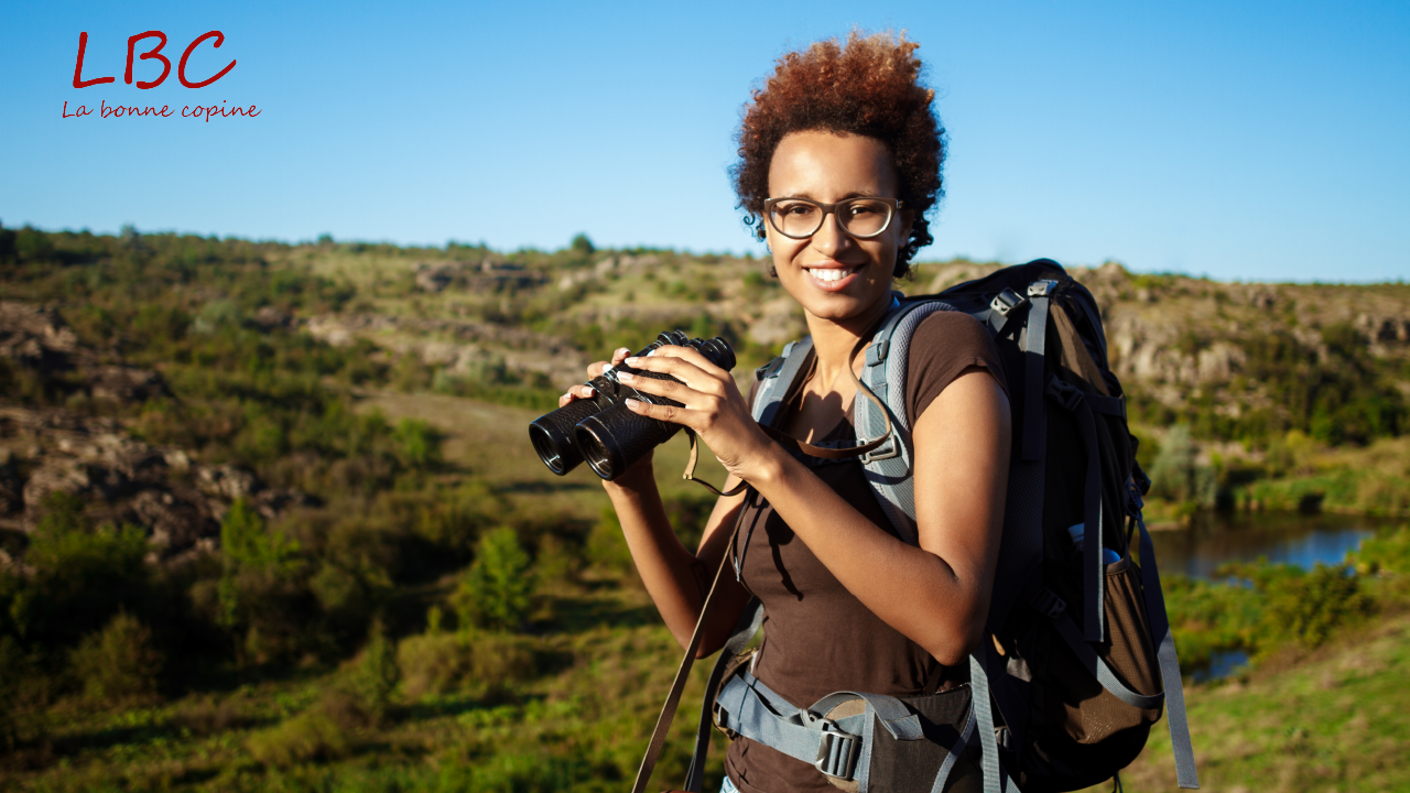 Smiling, equipped with her camera and backpack, a photographer enjoys a natural hilly landscape, ready to capture the beauty of the moment | labonnecopine.com/en
