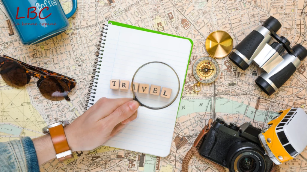 A hand holds a magnifying glass over the word ‘TRAVEL’ inscribed in a notebook, placed on a map. Around it, travel accessories suggest exploration and preparation for a new adventure | labonnecopine.com/en