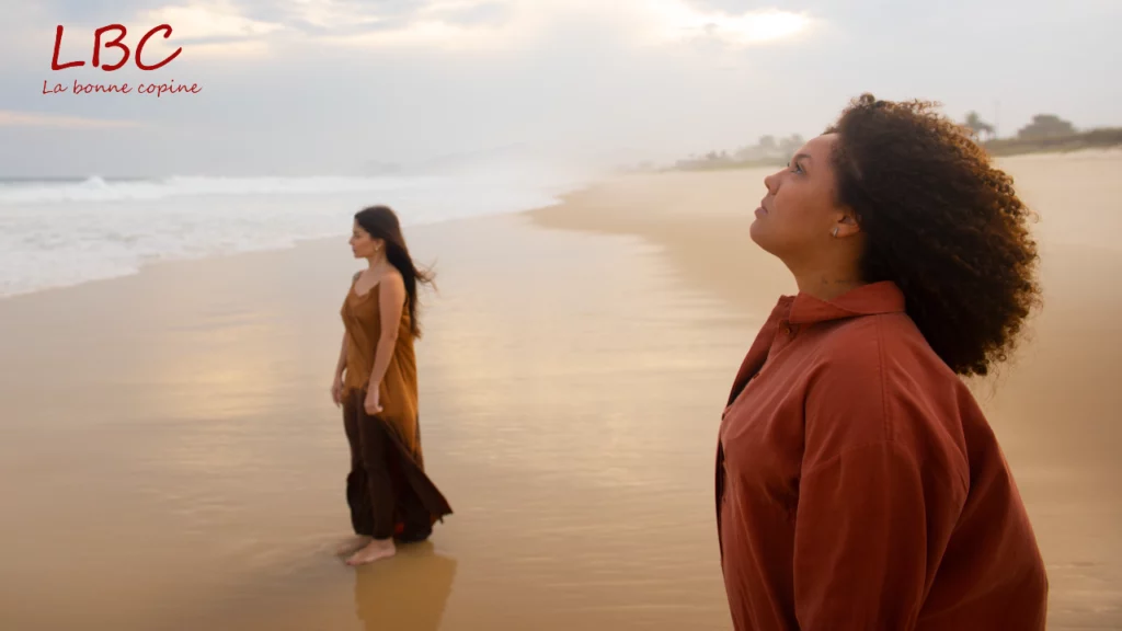 Two women walking silently on a calm beach; one gazes at the horizon, the other breathes deeply, both faces turned toward the sky, embodying mindfulness in nature.