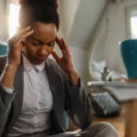 Assise au sol dans un intérieur apaisant, une femme en tailleur gris adopte une posture tendue, les mains posées sur les tempes. Son visage fermé trahit une surcharge mentale, symbole d’un quotidien sous pression | labonnecopine.com