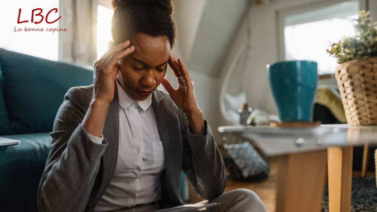 Assise au sol dans un intérieur apaisant, une femme en tailleur gris adopte une posture tendue, les mains posées sur les tempes. Son visage fermé trahit une surcharge mentale, symbole d’un quotidien sous pression | labonnecopine.com