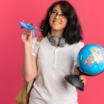Un sourire radieux éclaire le visage d’une jeune femme qui tient avec enthousiasme un avion miniature et un globe terrestre, symboles de ses rêves d’évasion | labonnecopine.com