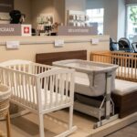 Intérieur d'un magasin de puériculture suisse affichant des couffins, berceaux et lits pour bébé, avec une femme enceinte qui regarde les produits. Panneaux de signalisation "COUFFINS ET BERCEAUX" et "SÉCURITÉ SUISSE". Prix visibles en CHF.