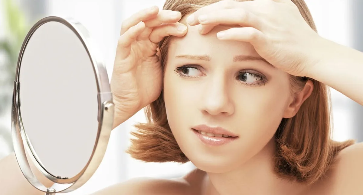 Une femme presse la peau de son front devant un miroir, geste à éviter.